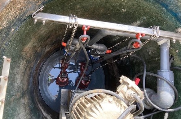 Overhead view of sewage pump suspended from metal bar with chains, connected to gray pipe and white air blower