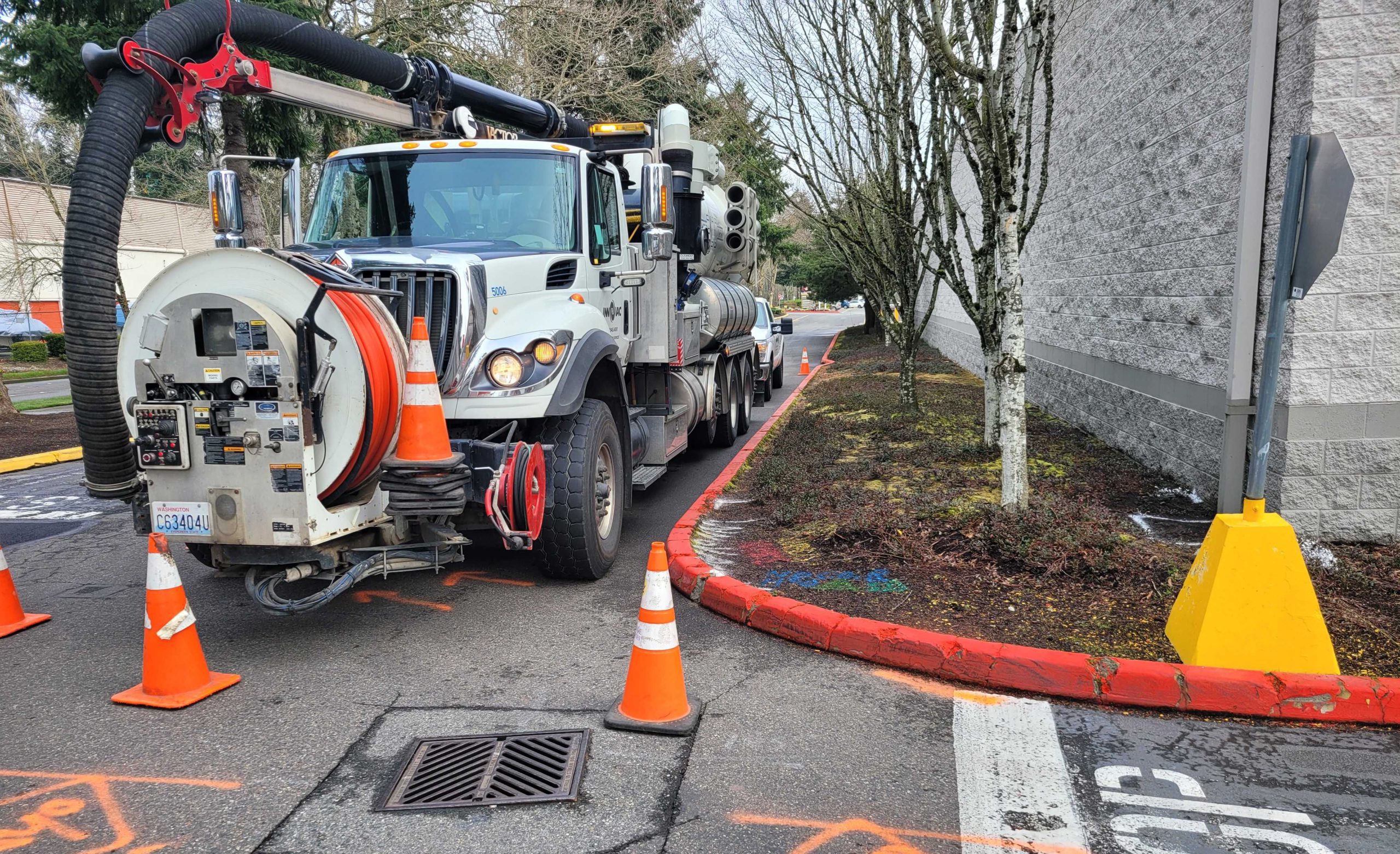 Truck with hydro vacuum, orange traffic cones and front-mounted hose spool parked at curb in front of storm drain grate