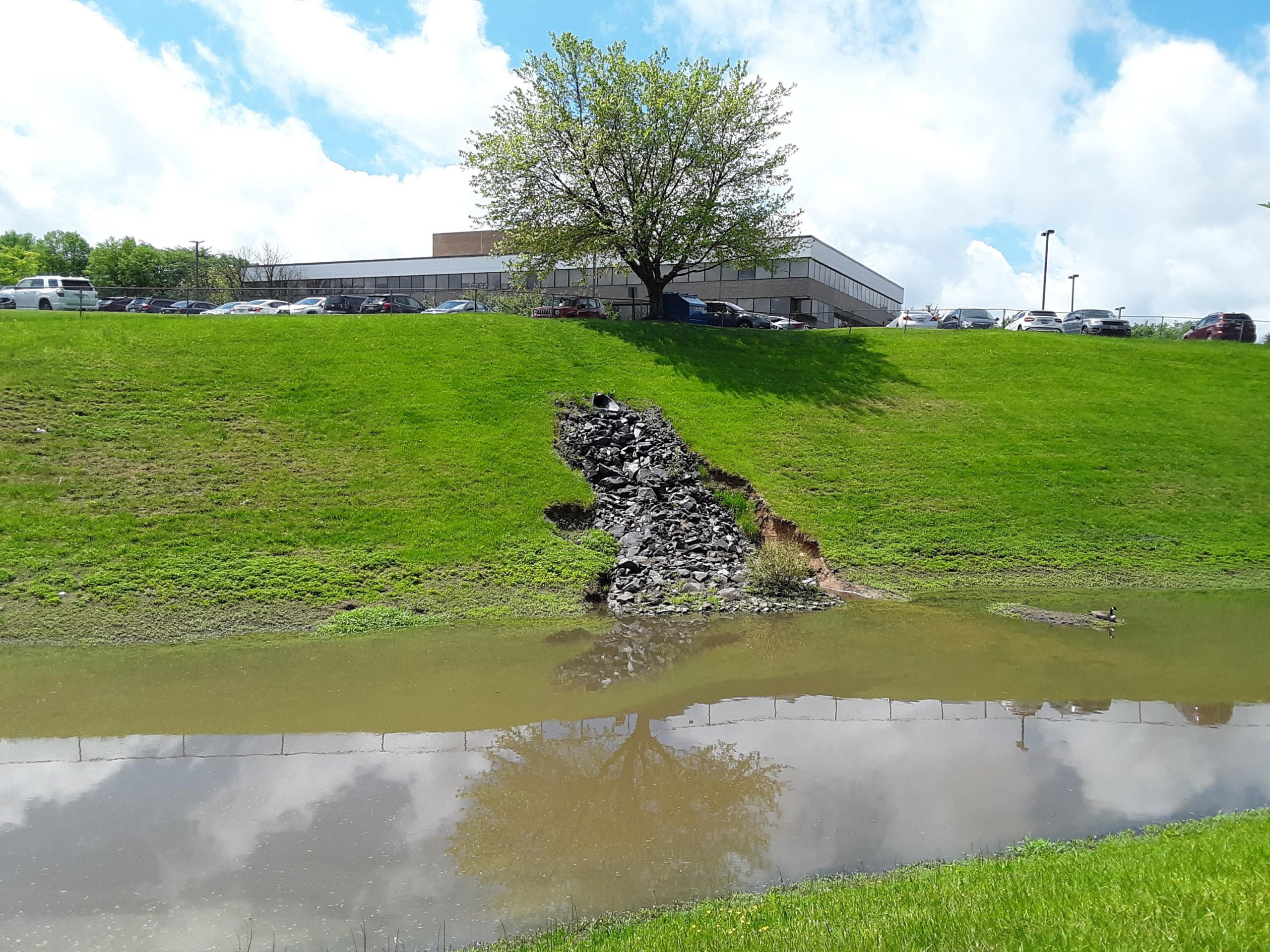 Stormwater detention basin at the foot of grassy hill split by a rock- filled gash below office building and parking lot