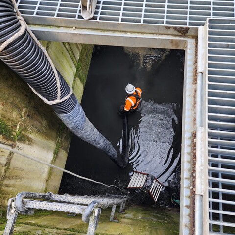 View down a street-level opening, past the grate, wall-mounted metal ladder and flexible pipe to construction worker standing in water at the bottom