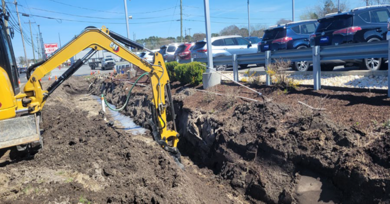 Excavator digging a trench alongside a full parking lot behind and below a metal barrier